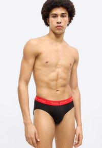 Μαύρα εσώρουχα με κόκκινη ζώνη μέσης που φέρει το λογότυπο "TOMMY HILFIGER". Το ύφασμα φαίνεται λείο και εφαρμόζει στενά, αναδεικνύοντας το σχήμα του σώματος.