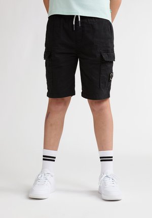 Schwarze Cargo-Shorts mit Taschen, Kordelzugbund, knöchellang, kombiniert mit weißen Sneakern und weißen Socken mit Streifen. Schlichter Hintergrund.