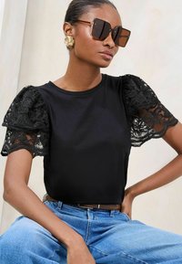 Black top with short lace puff sleeves; round neckline; paired with blue jeans. Accessories include oversized brown sunglasses and gold earrings.