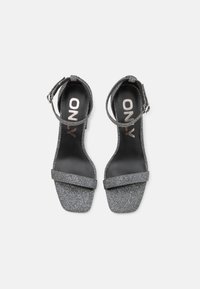 Grå glitrende sandaler med blokkhelt, med dobbel stroppdesign og ankelspenne, satt mot en svart innersåle.