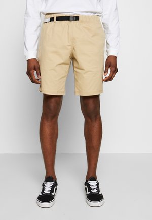 Homme portant un short beige avec une ceinture noire à boucle, un haut blanc à manches longues et des baskets noires et blanches, debout devant un fond uni.