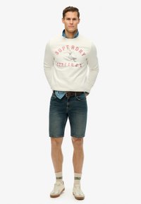 Felpa bianca con scritte e grafiche rosa, pantaloni shorts di jeans blu e sneakers beige con accenti marroni. Calzini leggeri con strisce verdi.