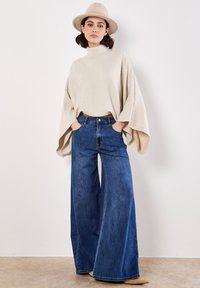 Maglione beige a costine con collo alto, abbinato a jeans in denim blu a gamba larga. La modella indossa un cappello di colore chiaro e posa con le mani in tasca.
