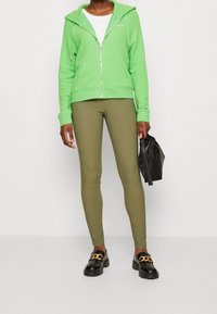 Sudadera verde con cremallera y bolsillos frontales, combinada con leggings oliva. Zapatos negros con detalles de cadena dorada y un pequeño bolso negro.