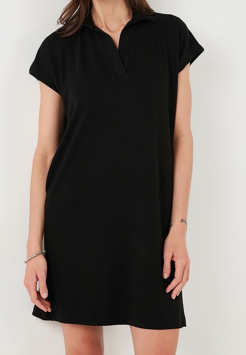 Femme portant une robe polo noire à manches courtes, debout devant un fond blanc uni, avec un bracelet et un petit tatouage au poignet visibles.