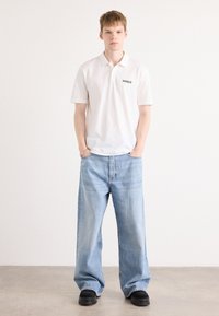 Hvit polo skjorte med svart "HUGO"-logo, kombinert med lyseblå vide jeans og svarte plattformsko. Enfarget bakgrunn. Enkelt design.