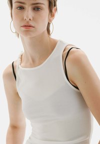 Top sin mangas blanco acanalado con un escote bajo, que presenta una tira retorcida en un hombro y tirantes ajustables negros por debajo.