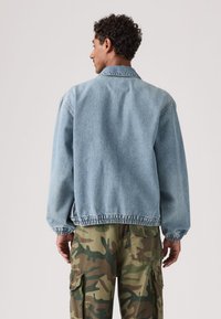 Φωτεινό μπλε τζιν μπουφάν με ελαστικές μανσέτες και μέση. Διαθέτει κλασικό γιακά και άνετη εφαρμογή, συνδυασμένο με παντελόνι καμουφλάζ cargo.