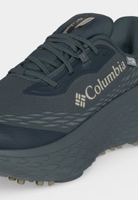 Σκούρο γκρι αδιάβροχο παπούτσι πεζοπορίας Columbia με υφή στη σόλα, κορδόνια και λογότυπο στο πλάι και στη γλώσσα, απεικονιζόμενο από μπροστινή πλευρική γωνία.