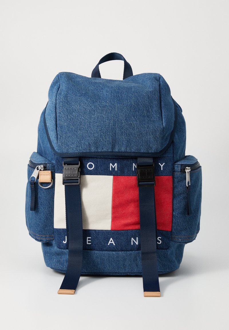 Denim backpack featuring a flap closure, navy straps, red, white, and blue logo detail, and exterior zip pockets for storage.