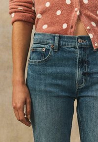 Siniset korkeavyötäröiset farkut, joissa on kuparinväriset yksityiskohdat ja hienovaraiset kulumat, yhdistettynä vaaleanpunaiseen pilkulliseen neuletakkiin.