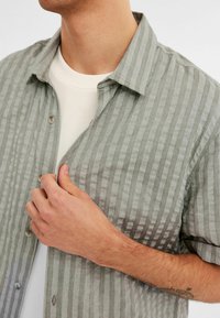Kurzärmliges Hemd in Hellgrau mit vertikalen Streifen, entspanntem Kragen und strukturiertem Stoff. Über einem weißen Shirt getragen.