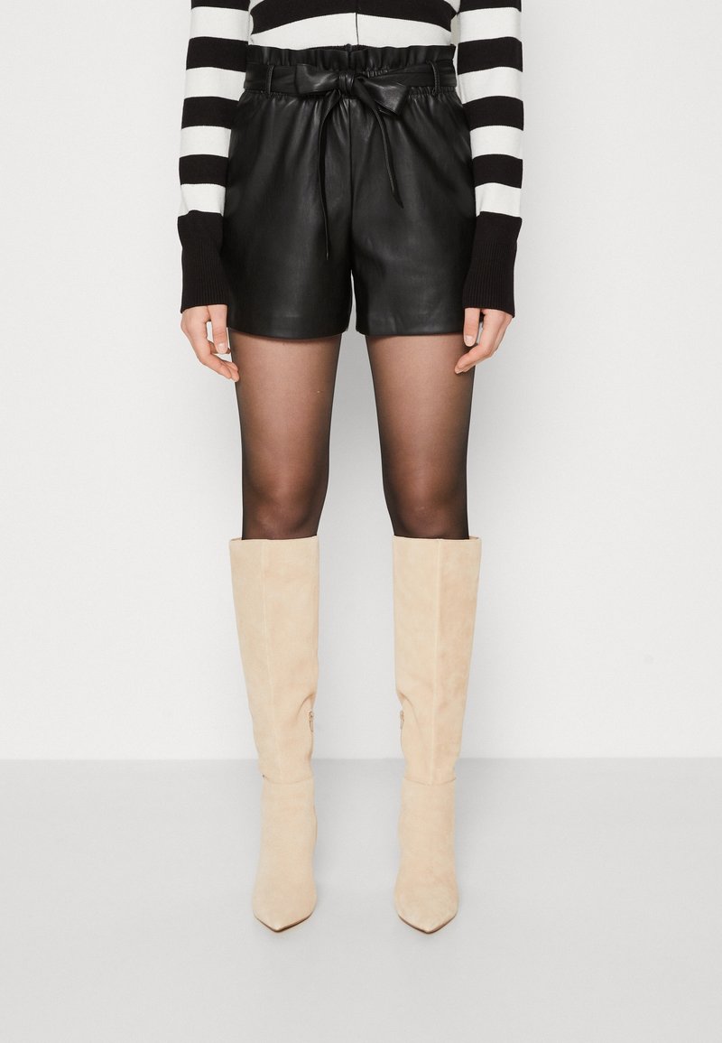 Personne portant un short en cuir noir avec une ceinture nouée en noeud, des collants noirs transparents, des bottes en daim beige montant jusqu'aux genoux et un pull à rayures noires et blanches.