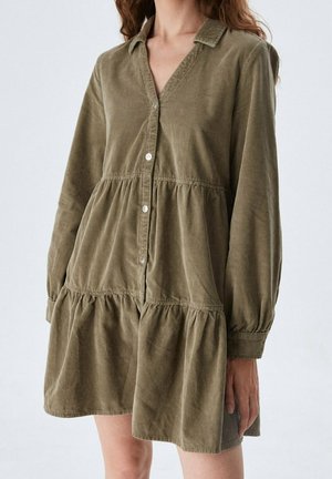 Vrouw draagt een lange mouw, olijfgroene, knoopsluitende, gelaagde jurk met kraag en staat tegen een effen lichte achtergrond.