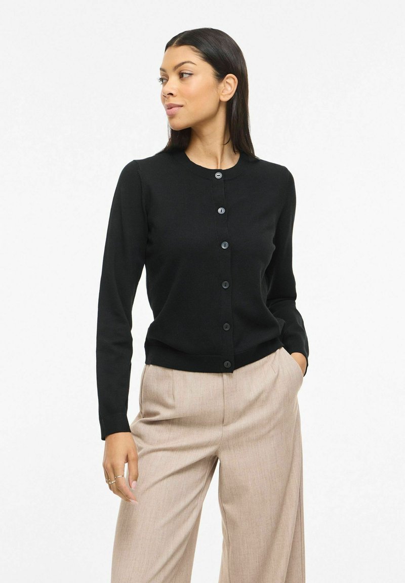 Vrouw met een zwart geknoopt vest en beige broek, staand met één hand in de zak, kijkend naar de zijkant tegen een witte achtergrond.