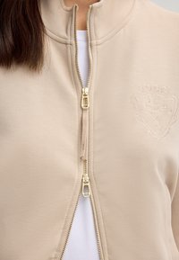 Giacca beige con zip e colletto alto, zip dorata e logo ricamato. Tessuto morbido con una consistenza liscia, caratterizzato da uno strato interno bianco.