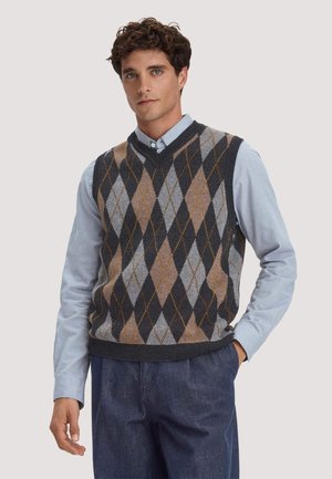 Junger Mann, der ein blaues Hemd mit Knöpfen und eine dunkle ärmellose Argyle-Weste mit blauen Hosen trägt.