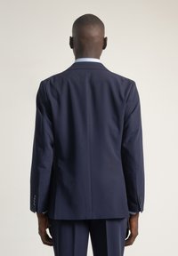 Blazer entallado azul marino con textura suave, solapas amplias y puños abotonados, visto desde atrás sobre un fondo liso.