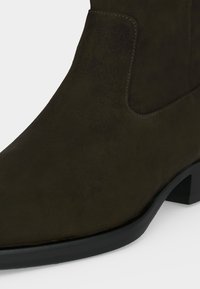 Gros plan d'une botte en daim brun foncé avec un talon bas noir et un détail de couture sur le panneau latéral élastique.