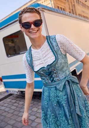 Jonge vrouw die glimlacht, met een zonnebril en een blauw geborduurd dirndljurk met een witte kanten blouse, staand op een straat met kasseien.