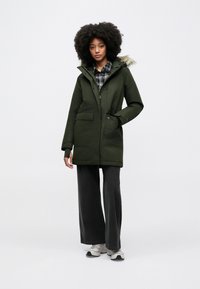 Olijfgroene parka met een capuchon gevoerd met nepbont, voorzakken en een rits. Gecombineerd met donkere, wijd uitlopende broekspijpen en witte sneakers.