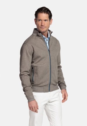 Man wearing light brown zip-up jacket over blue shirt and white pants, standing against plain light background, looking left.