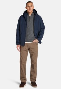 Modelo masculino de pie, vistiendo una chaqueta azul marino sobre un suéter gris y pantalones color beige, con zapatos oscuros. El fondo neutral enfatiza el atuendo.