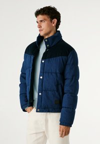 Temno modra puffer jakna s kordurojnimi detajli, zatiči in elastičnimi manšeti. Ima prešito teksturo in ravno obrobo.