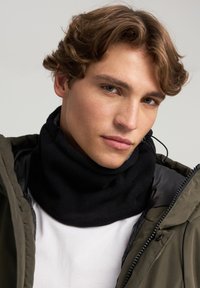Giovane uomo con capelli castani mossi che indossa un colletto nero, una camicia bianca e una giacca verde oliva su uno sfondo neutro.