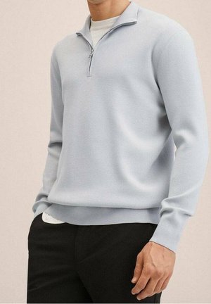 Hellblauer Pullover aus weichem Strickmaterial, mit einem halbseitigen Reißverschlusskragen, gerippten Bündchen und Saum. Über einem weißen Hemd getragen.
