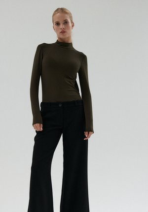 Młoda kobieta o blond włosach nosi ciemnozielony top z długim rękawem oraz czarne, szerokie spodnie z wysokim stanem, stoi na jednolitym tle.