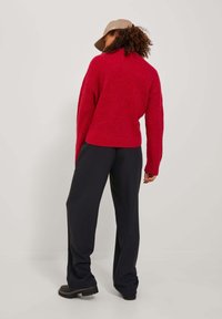 Röd räfflad tröja med hög krage, matchad med högmidjade svarta vida byxor och kraftiga svarta stövlar. Naturliga lockar och en keps synlig.