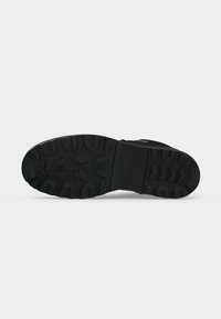 Semelle de botte noire avec des rainures profondes et le logo Tommy Hilfiger visible au centre sur un fond gris clair uni.