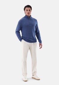 Blauer Strickpullover mit einem Reißverschlusskragen, kombiniert mit cremefarbenen Plisseehosen und hellbeigen Sneakers, vor einem schlichten Hintergrund stehend.