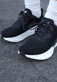 Baskets de sport noires avec dessus texturé, semelle blanche et lacets réfléchissants. Dispose d'un design épuré et d'un soutien rembourré.
