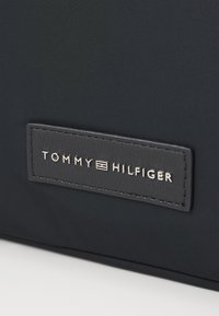 Czarny materiał nylonowy z prostokątną skórzaną naszywką z wytłoczonym logo "TOMMY HILFIGER" w srebrnym kolorze.