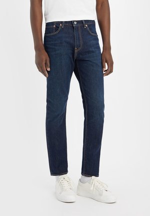Mann trägt dunkelblaue Slim-Fit-Jeans, weiße Sneakers und ein weißes Hemd und steht vor einem schlichten weißen Hintergrund.
