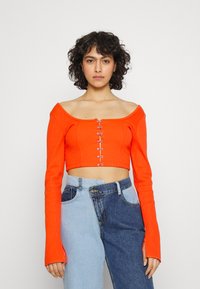 Sweater cropped laranja com design de ombro caído, textura canelada e detalhes em gancho na parte da frente. Combinado com jeans de denim azul assimétricos.