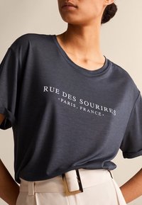 Dunkelgraues T-Shirt aus glattem Stoff, mit dem Aufdruck "RUE DES SOURIRES - PARIS, FRANKREICH" in weißer Schrift auf der Vorderseite. Kurzärmlig.
