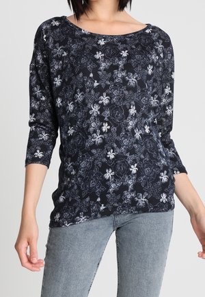 Vrouw draagt een zwart shirt met lange mouwen met witte bloemmotieven en lichtblauwe spijkerbroek tegen een effen lichte achtergrond.