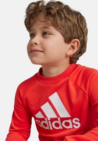 Sudadera roja con cuello redondo de canalé y logo blanco de Adidas. Presenta mangas raglán y una textura de tejido suave.