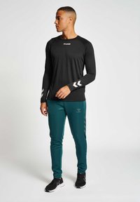 Schwarzes Langarmshirt mit weißem Logo und Chevron-Designs. Türkise Hose mit dezentem Logo und hellgrauen Streifen. Schwarze Sportschuhe.