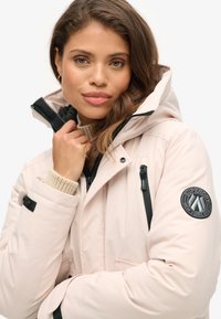 Jasnoróżowa kurtka z kapturem, z czarnymi akcentami, zamkami błyskawicznymi i naszywką z logo. Posiada teksturowany ściągacz, wielofunkcyjny design z wieloma kieszeniami oraz materiał odporny na wodę.