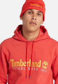 Sudadera de algodón roja con el logo y texto amarillo de Timberland, con cordones y un cuello acanalado. Combinada con un gorro de punto rojo a juego.