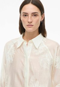 Blouse crème légère et translucide avec broderies florales blanches, boutonnage à l'avant et col classique. Texture lisse et coupe décontractée.