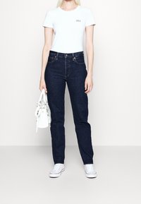 Woman wearing a white GAP t-shirt, dark blue jeans, white sneakers, holding a white handbag against a plain light background.