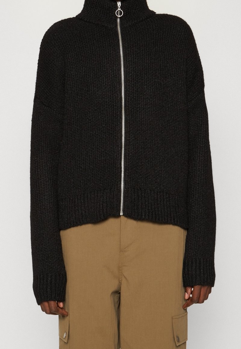 Suéter negro de punto corto con cuello alto, cremallera frontal plateada y dobladillos acanalados. Presenta mangas anchas y tejido texturizado.