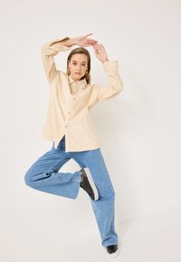 Camicia beige con bottoni e colletto, abbinata a jeans a gamba larga blu chiaro e sneakers nere. La camicia ha una vestibilità comoda e maniche lunghe.