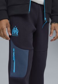 Marineblauwe trainingsbroek met een blauw logo en tekst, voorzien van een zijzak met rits in een lichterblauwe stof en een elastische tailleband.