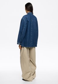 Giacca in denim blu scuro con una vestibilità morbida, caratterizzata da maniche lunghe, colletto classico e cuciture a vista. Abbinata a pantaloni larghi beige.